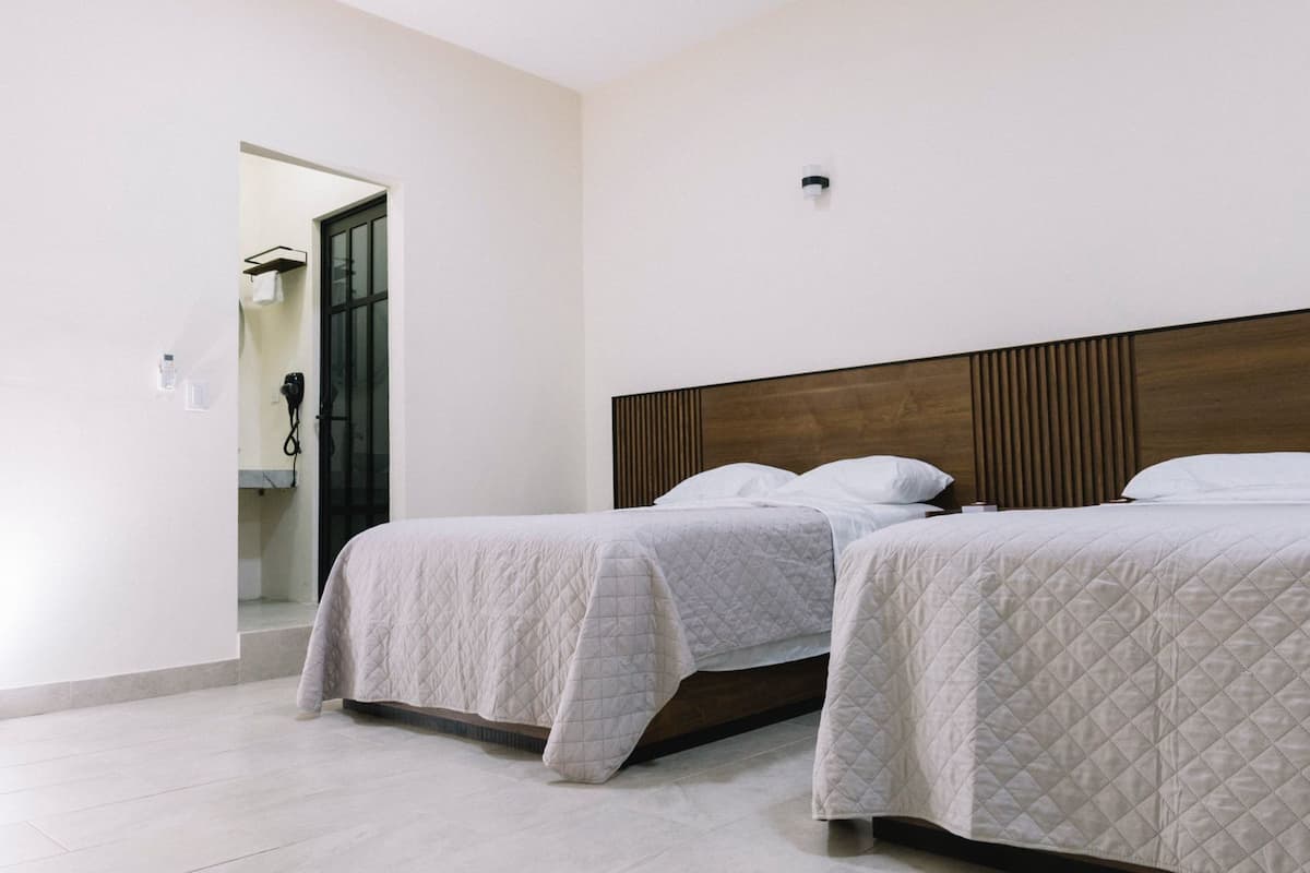 Habitación de hotel con dos camas, cabecero de madera y un baño pequeño a la vista a través de una puerta abierta.