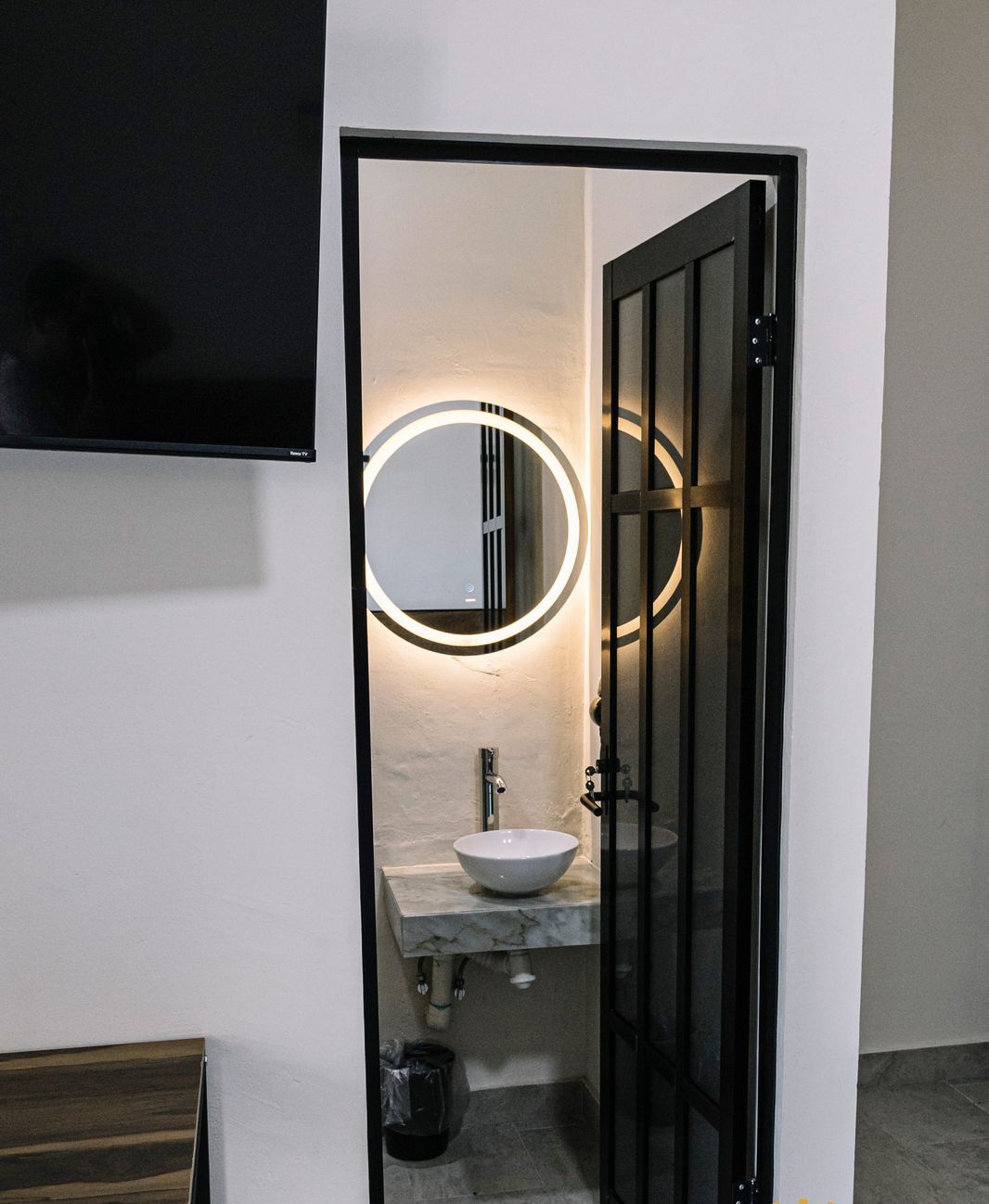 Un baño moderno con un lavamanos blanco, un espejo redondo con iluminación y una puerta negra.