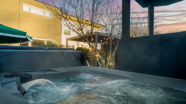 A hot tub with a waterfall feature, surrounded by a covered seating area and a building with large windows in the background. - The Happy Hideaway (Seattle)