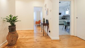 A hallway with wooden flooring, a rustic vase with green plants, a small table with a vase, and a kitchen visible through an open door.
