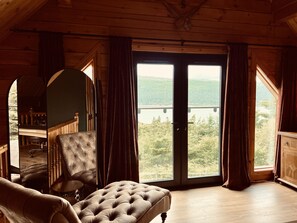 A wooden cabin with a large sliding glass door leading to a balcony with a scenic view of a lake and forest. Inside, there is a tufted sofa and a chair.