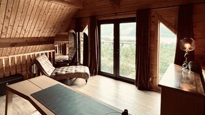 Ein Zimmer in einer Holzhütte mit Schlafkoje, Schreibtisch mit Lampe und Blick auf eine bewaldete Landschaft durch große Fenster.