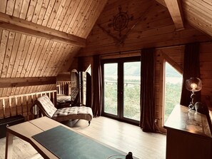 A wooden cabin room with a loft bed, a desk with a lamp, and a view of a forested landscape through large windows.