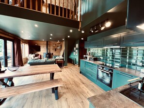 Een moderne keuken met blauwgroene kastjes, een houten eettafel en een gezellig zithoek met een bank en open haard.