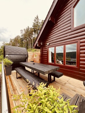A wooden cabin with a deck, a barbecue grill, and a large window offering a view of the surrounding forest.