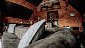 A cozy cabin interior with a wooden ceiling, a sofa with cushions, a framed deer picture, and a lamp.