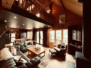 A spacious living area with a high ceiling, wooden beams, a large window offering a view of the outdoors, a dining table, and a sofa.