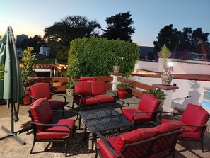 An outdoor patio with red chairs and tables, surrounded by potted plants and a green hedge.
