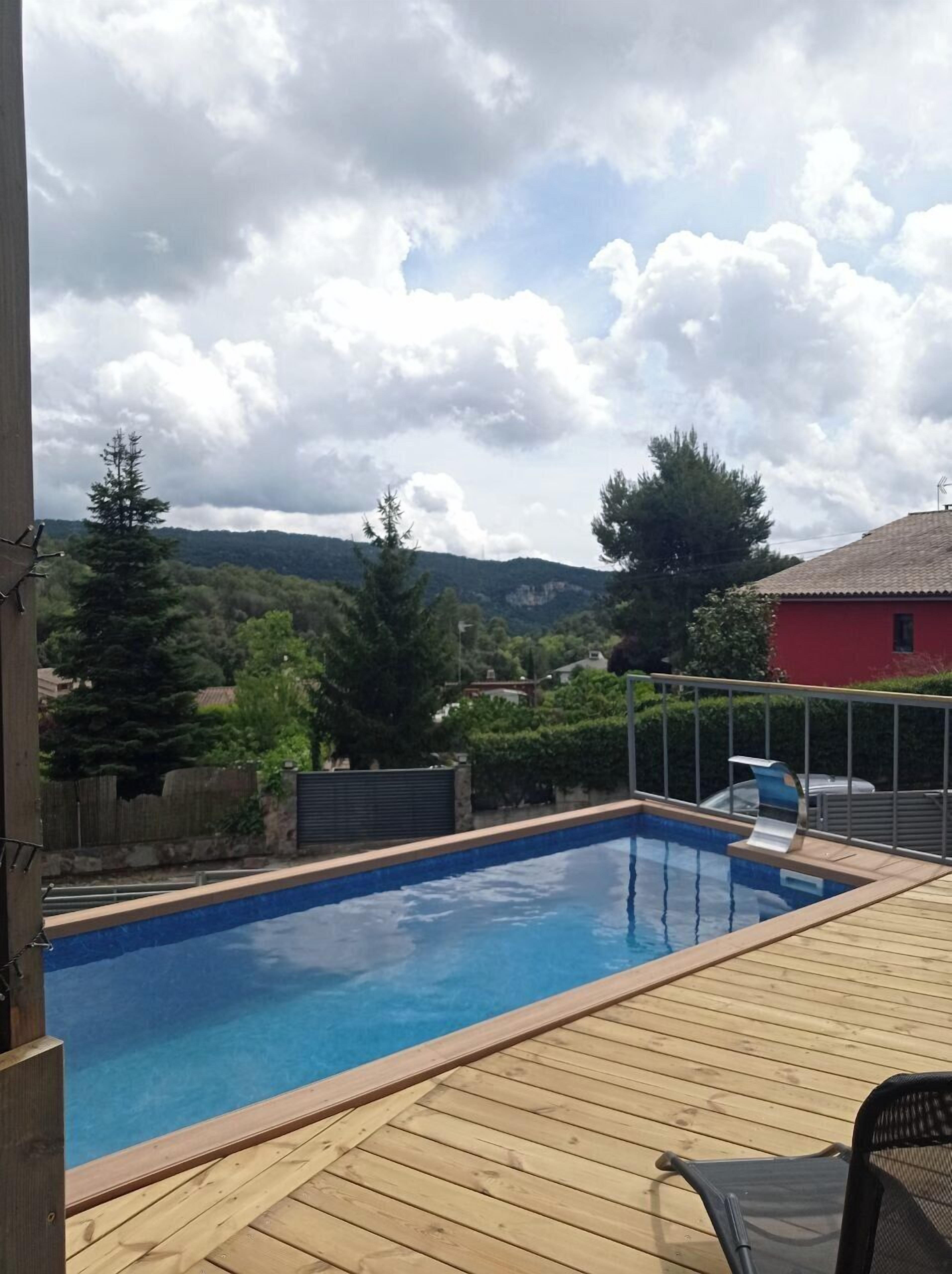 Ein Schwimmbad mit Holzterrasse, umgeben von viel Grün und einem roten Gebäude im Hintergrund.