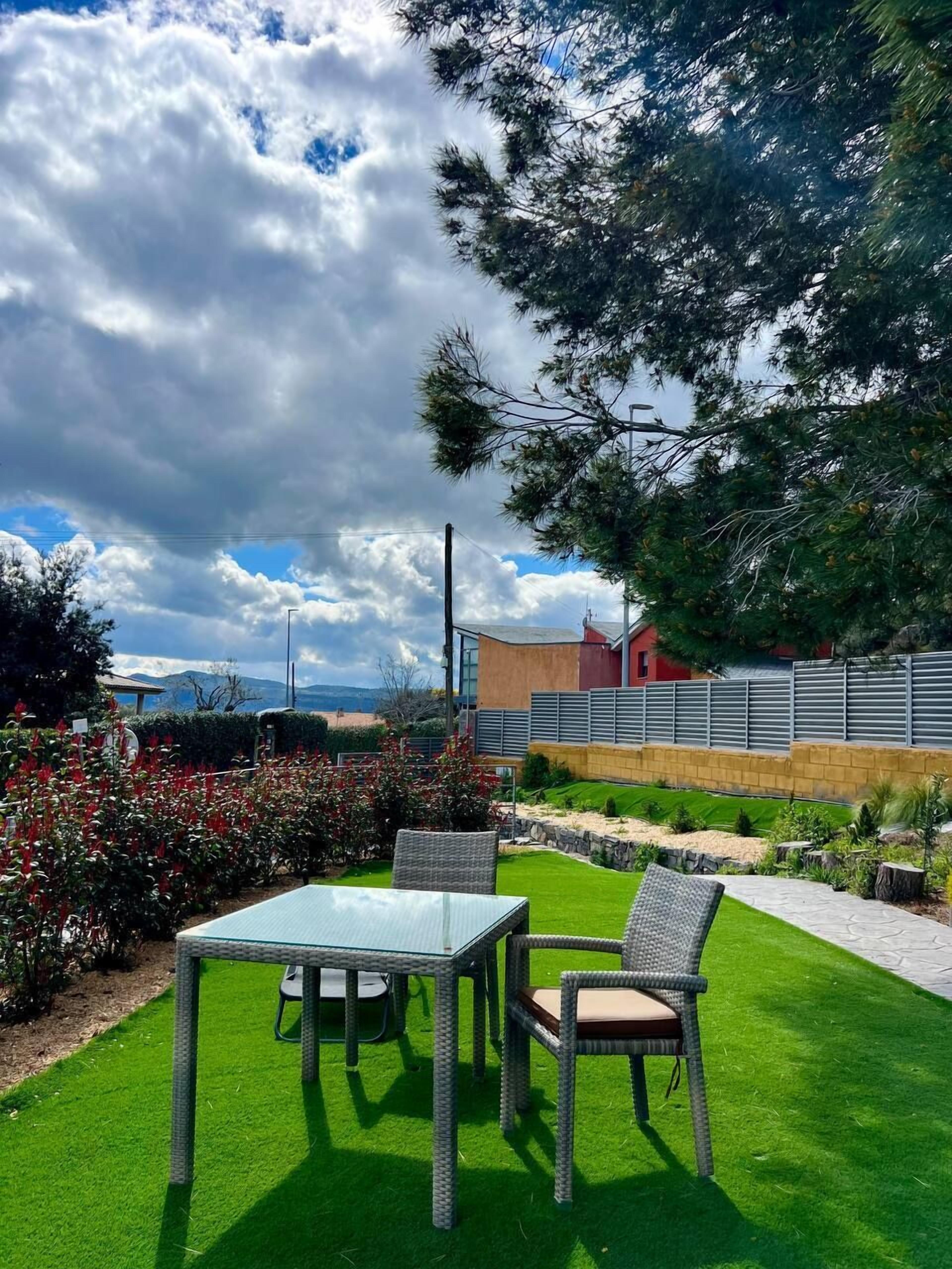 A patio area with a table and chairs, surrounded by greenery and a view of a building in the background.