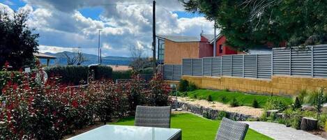 A patio area with a table and chairs, surrounded by greenery and a view of a building in the background.