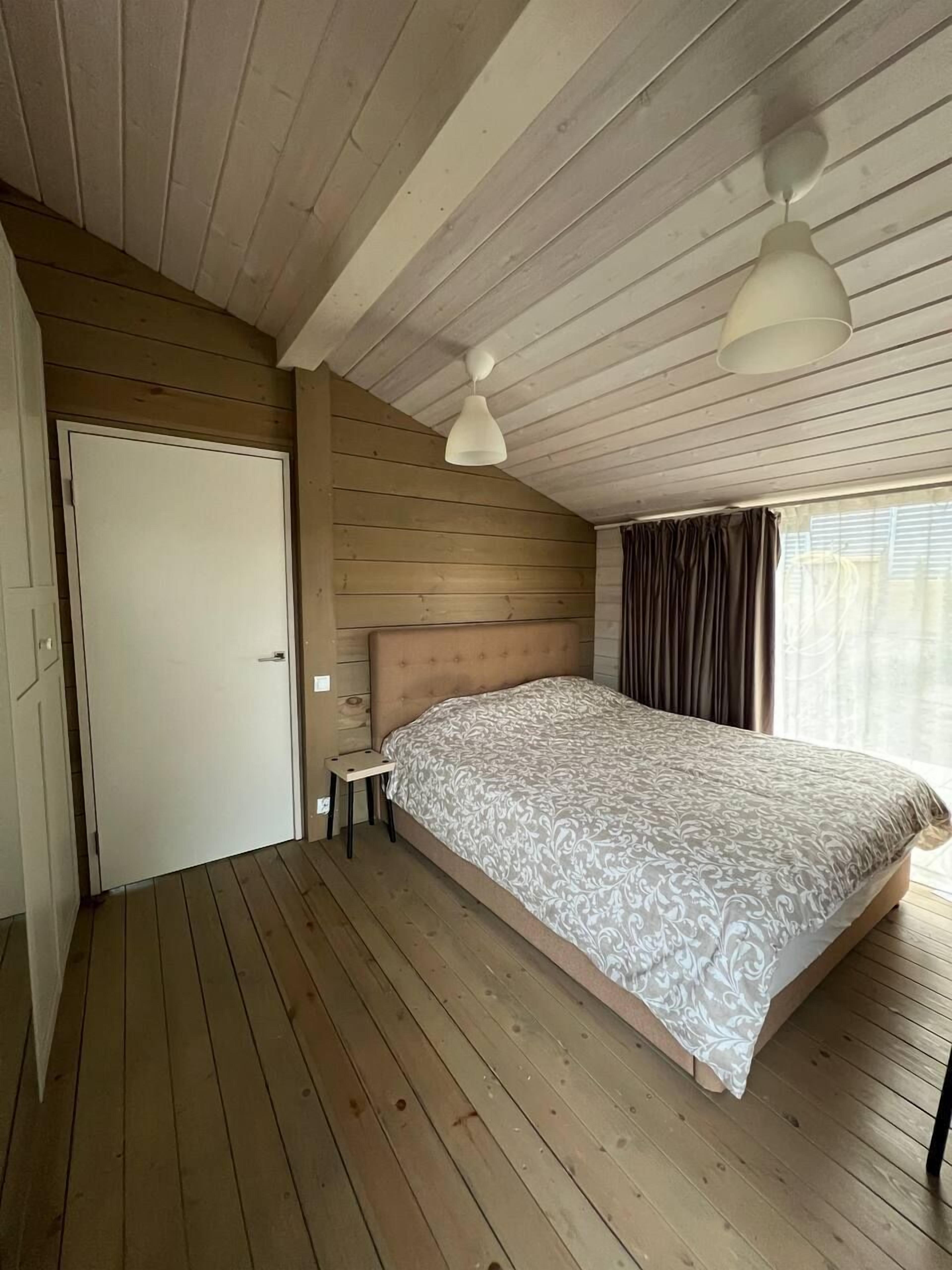Un dormitorio con piso de madera, una cama con colcha estampada, una mesita de noche pequeña, una puerta blanca y una lámpara en el techo.