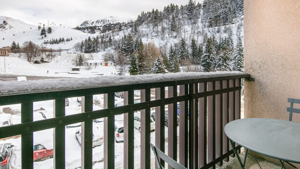 Un balcon doté d’une rambarde en métal, donnant sur un paysage enneigé où l’on aperçoit des voitures garées et, au loin, un télésiège.