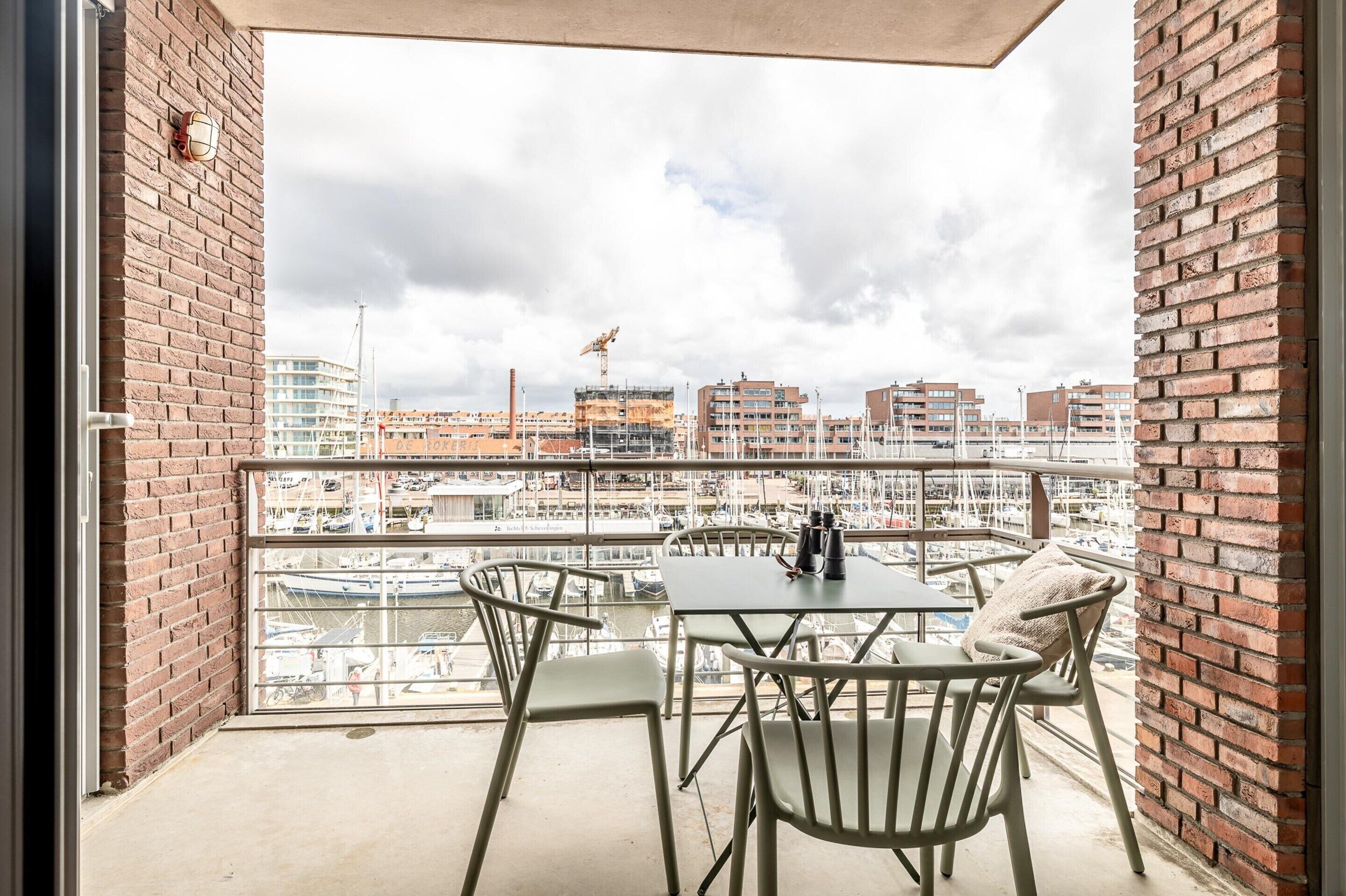 A balcony with a table and chairs, overlooking a marina with boats and buildings in the background.
