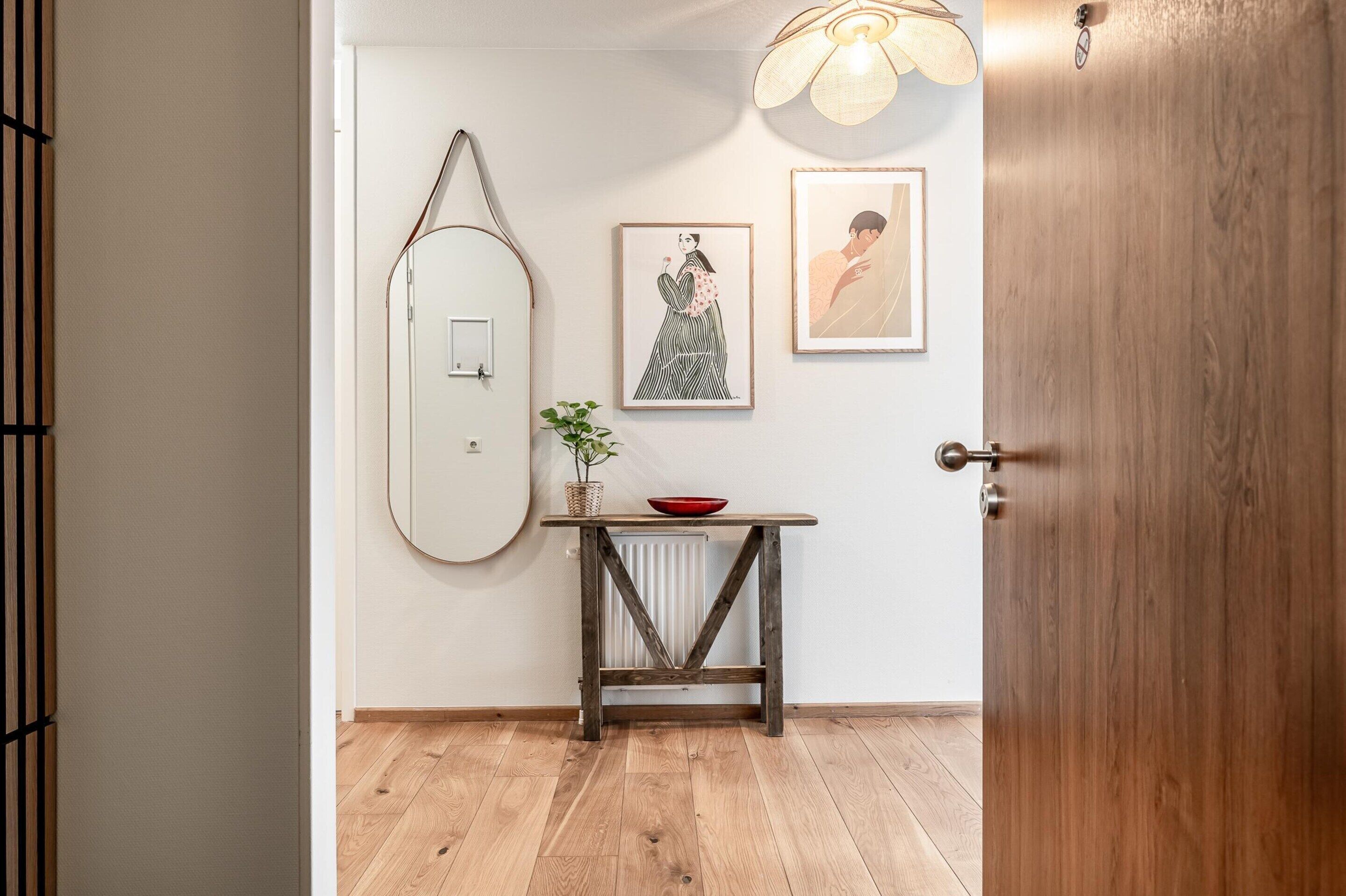 Een gang met een houten vloer, een witte deur, een houten tafel met een rode schaal, en twee ingelijste foto's aan de muur.