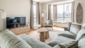 Ein modernes Wohnzimmer mit Flachbildfernseher, einer Couch, einem Sessel, einem Couchtisch und einem großen Fenster mit Blick auf die Stadt.