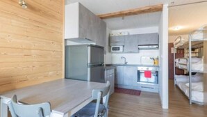 Eine kleine Küche mit Kühlschrank, Mikrowelle und Spüle. Ein Esstisch mit Stühlen und ein Etagenbett mit weißer Bettwäsche sind vorhanden.