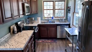 Una cocina con armarios de madera oscura, encimeras de granito, refrigerador de acero inoxidable, microondas, tostadora y ventana con vista a árboles.
