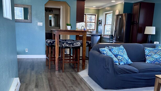 Un salón con un sofá azul oscuro, cojines estampados, una mesa de centro de madera y una cocina al fondo.