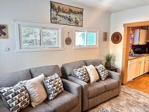 A living room with two grey sofas, patterned cushions, a painting above the windows, and a kitchen area visible through an open door. - Auke Ridge Retreat - a short walk to the harbor! (Juneau)
