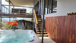A modern outdoor hot tub area with a wooden deck, a staircase, and a view of a building in the background.