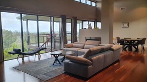 A spacious living room with a large sectional sofa, a dining table with chairs, and a balcony with a view of the forest.