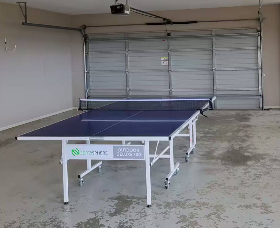 A ping pong table labeled 'TerraSphere Outdoor Deluxe 700' in a garage with a concrete floor and a closed garage door in the background.