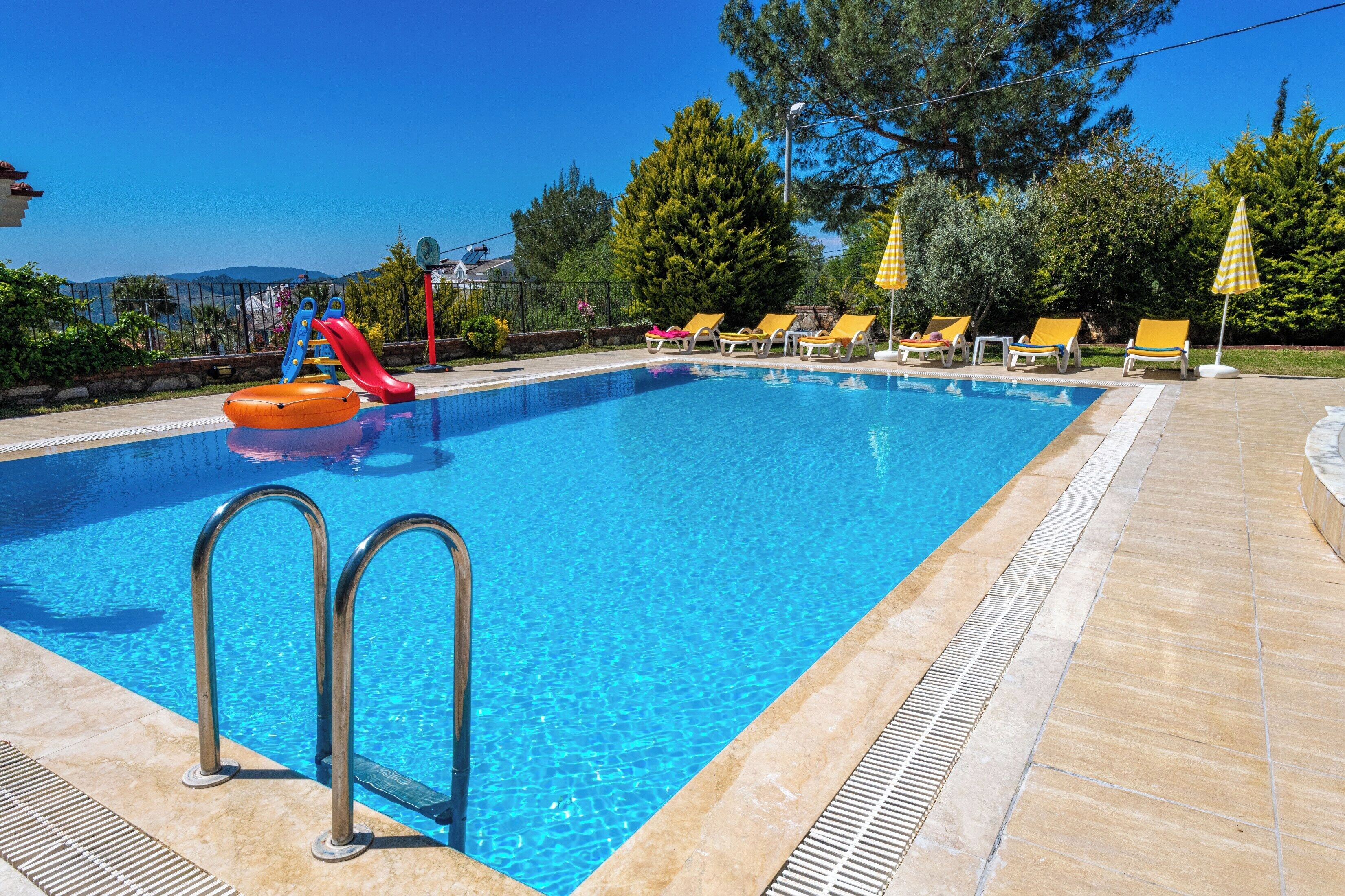 A swimming pool with a slide, sun loungers, and a view of trees and mountains.