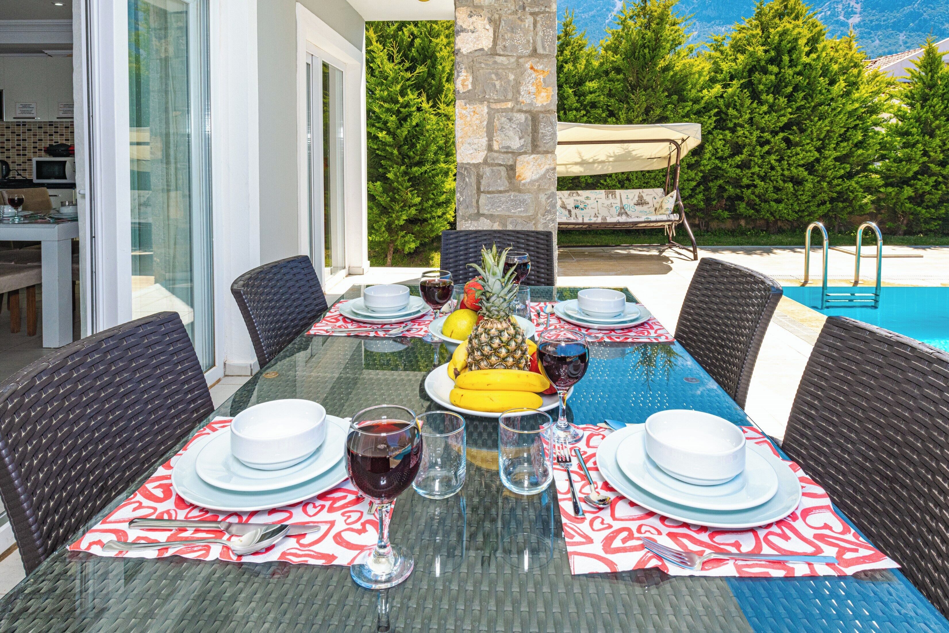 A dining table set for six with a tropical fruit centerpiece, a pool in the background, and a stone pillar.