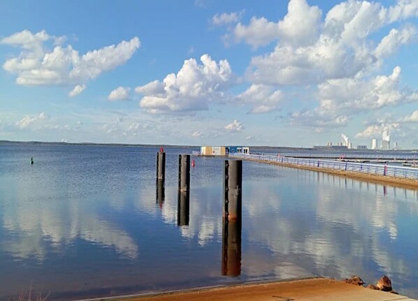 A calm waterfront with a pier extending into the water, a distant industrial facility, and a clear sky. - Floating House at Lake Barwalder See (Boxberg/O.L.)