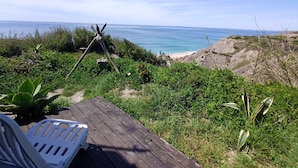 Une terrasse en bois avec une chaise en plastique blanche, donnant sur une plage au terrain rocheux et offrant une vue dégagée sur l’océan.