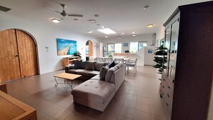 A spacious living area with a sofa, a coffee table, and a dining table with chairs. There is a wooden door and a large window providing natural light. - SandNSea - Only Steps to the beach -Pet Friendly Accommodation (Burrum Heads)