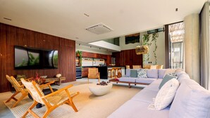 A modern living room with a large TV, wooden furniture, a sofa, and a kitchen in the background. - Casa Alma - By Living Milagres (Passo de Camaragibe)