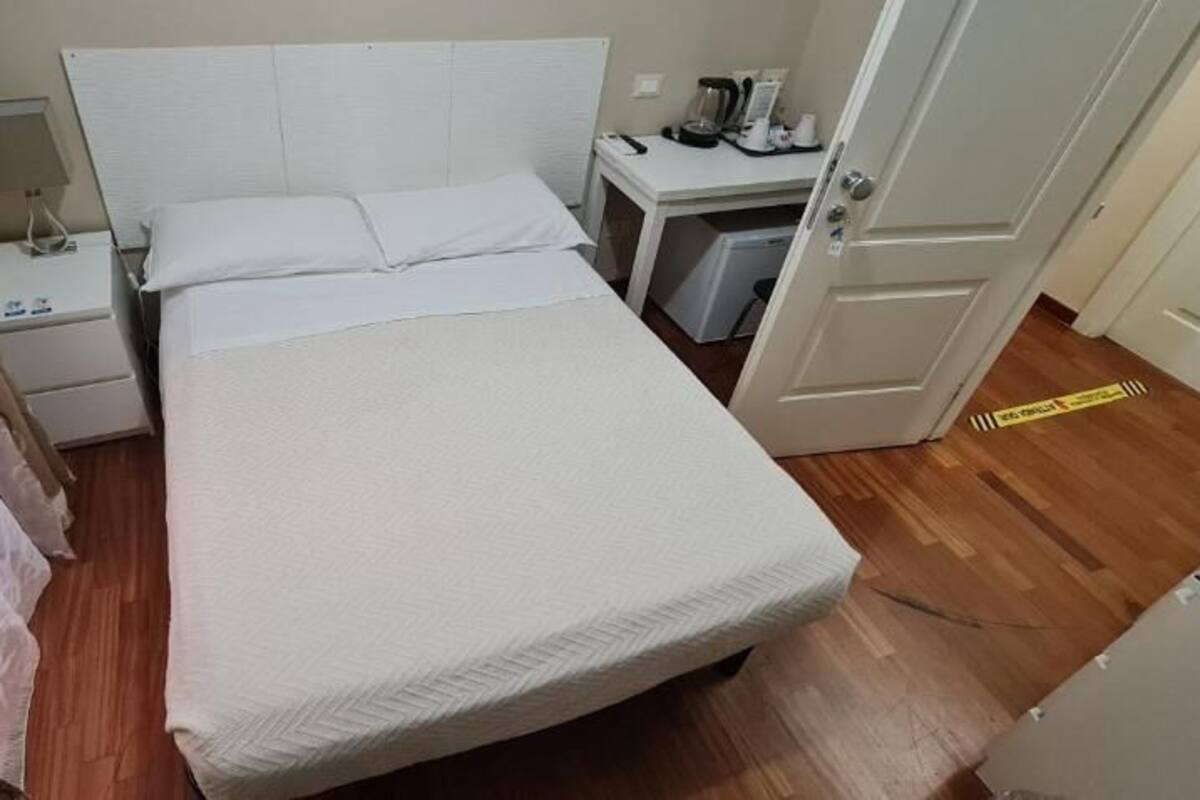 Un lit simple avec un matelas et des oreillers blancs, une table de chevet avec une lampe, un petit bureau avec une chaise et une porte ornée d’un ruban de signalisation jaune.