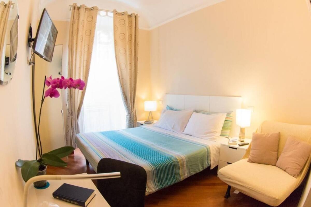 Une chambre d’hôtel comprenant un lit, des tables de chevet, un bureau avec une chaise, une petite table avec une plante et un miroir au mur.