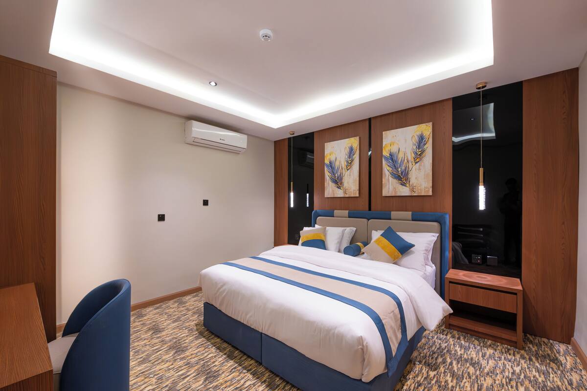 A hotel room with a bed, a desk, a chair, a nightstand, and artwork on the wall.
