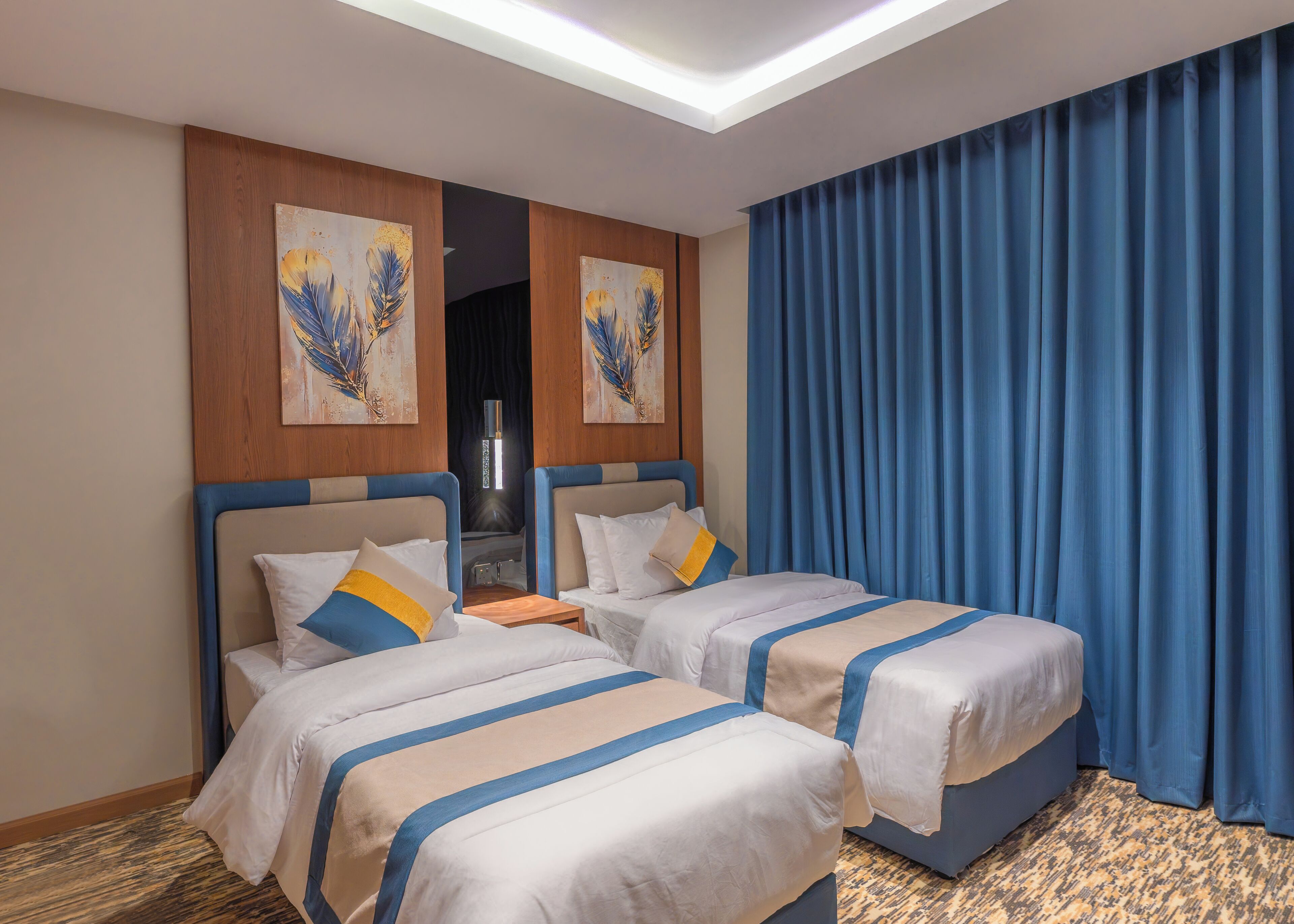Une chambre d’hôtel avec deux lits, une tête de lit en bois, une grande fenêtre avec des rideaux bleus et des œuvres d’art accrochées aux murs.