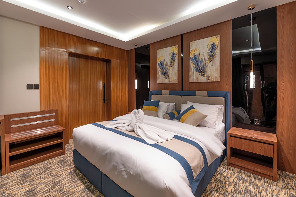 A hotel room with a large bed, wooden headboard, a bench, a small table, and artwork on the walls.