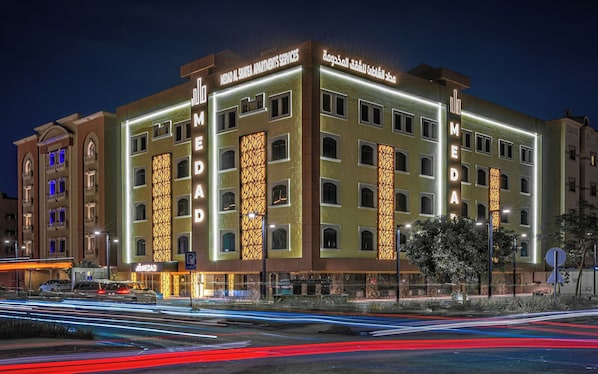 A multi-story hotel with illuminated signage, a parking sign, and a view of the street with light trails. - MEDAD ALSHATEA SERVICED APARTMENTS (Dammam)