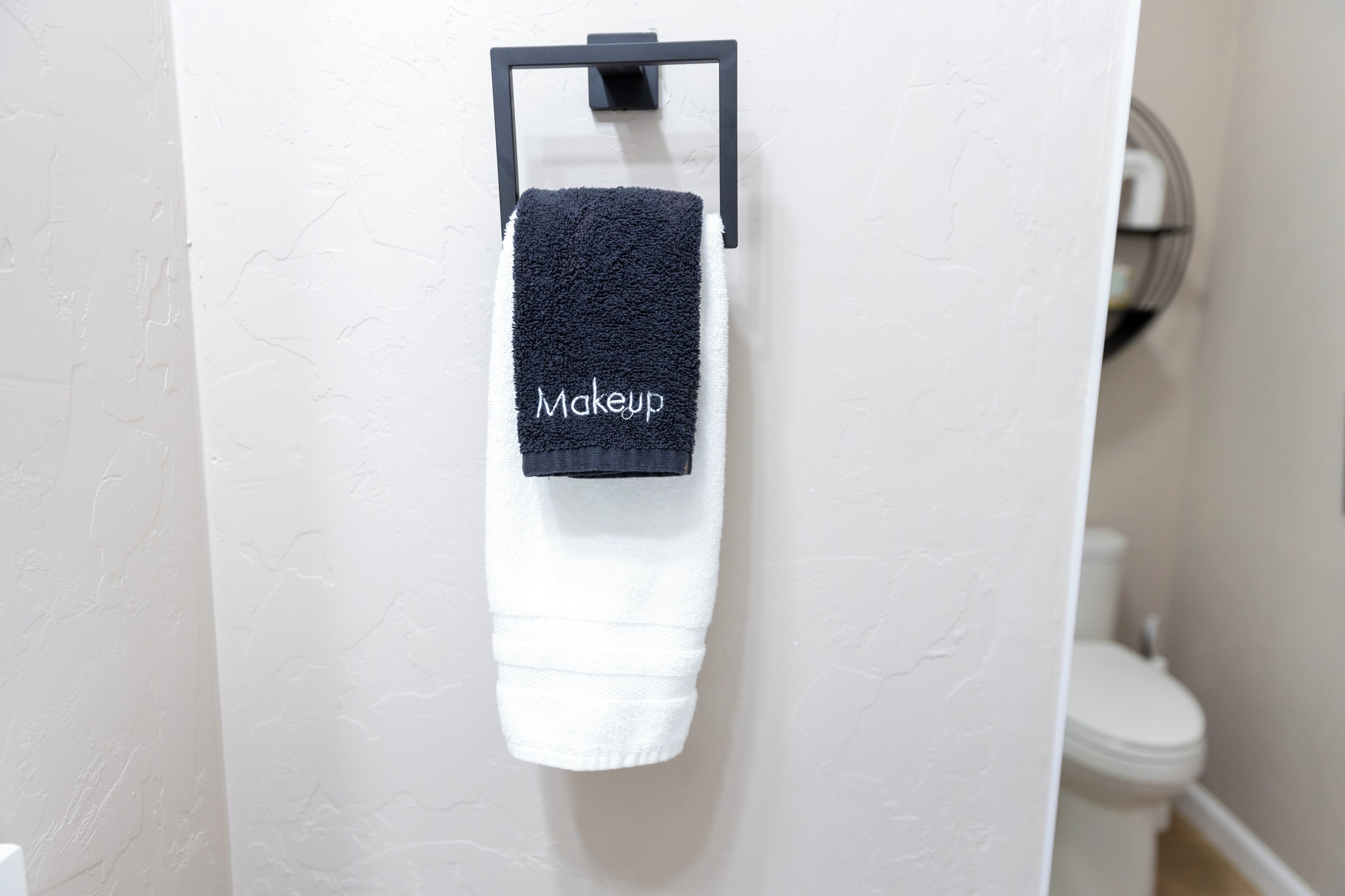 A white towel with a black tag labeled "Makeup" hanging on a black towel rack in a bathroom.