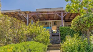 A building with a covered entrance, a staircase leading to the main door, and a garden area with various plants. - Casa Libecciu (Lumio)