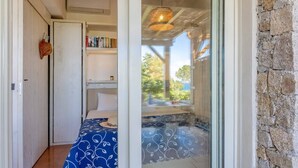 A room with a bed, a small table, a shelf with books, and a view of the outdoors through a glass door. - Casa Libecciu (Lumio)