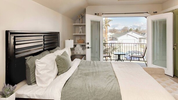 Une chambre à coucher comprenant un lit, des tables de chevet, une commode, une fenêtre et un balcon avec vue sur les maisons.