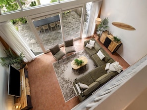 Una sala de estar moderna con sofá, mesa de centro y televisor, un balcón con comedor, y un mueble de madera con plantas.