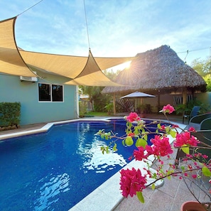 A swimming pool with a thatched-roof structure, a shaded area, and blooming bougainvillea.