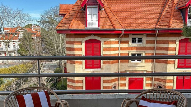 A balcony with wicker chairs and red-striped cushions, overlooking a building with a red-tiled roof and striped facade.