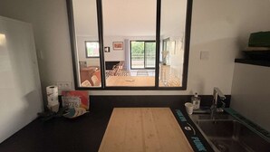 A kitchen with a large window, a wooden cutting board, a sink, and a dining area visible through the window.