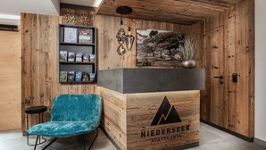 A reception desk with a wooden finish and a logo, a blue velvet chair, a bookshelf with magazines, and a framed picture on the wall. - Apartments Niederseer - Reiterkogel (Saalbach)