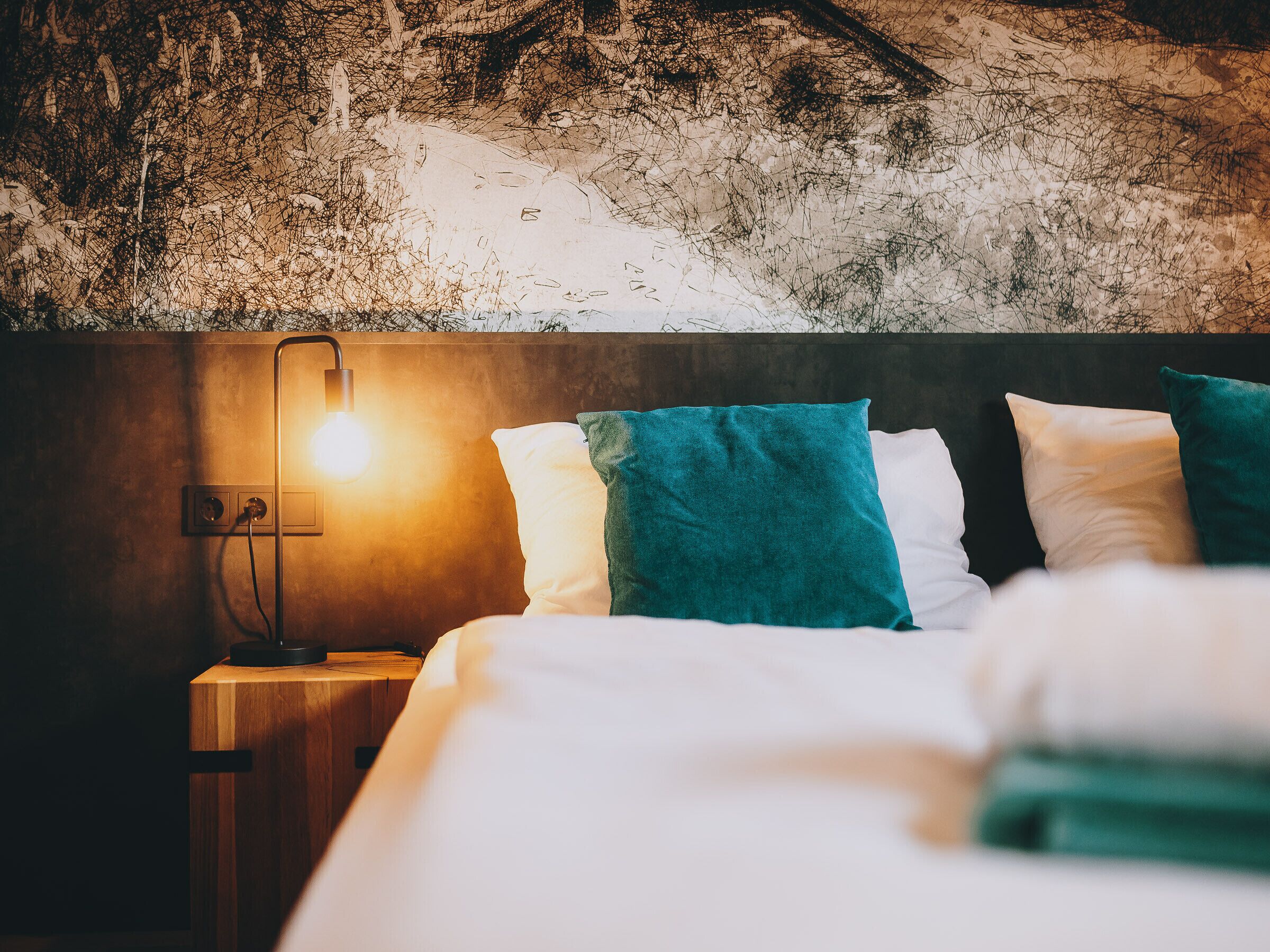 Un letto rifatto con cuscini bianchi e color acquamarina, un comodino in legno con una lampada e una parete testurizzata alle spalle.