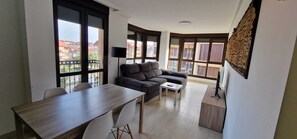 A modern living room with a sofa, a dining table, and large windows offering a view of buildings. - Apartamentos Gran Socaire de Noja (Noja)